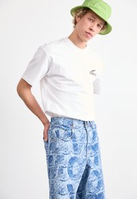 T-shirt en coton blanc avec un logo sur le devant, associé à un jean bleu avec un motif texturé. Un chapeau seau vert clair complète le look.