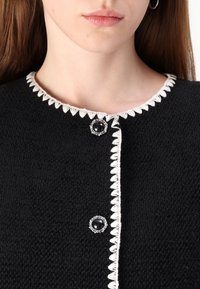 Primo piano di un cardigan nero con texture, rifinito con bordo bianco a forma di conchiglia e due bottoni decorativi neri e argentati vicino al colletto e al petto.