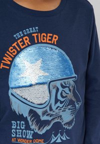 Granatowa koszulka z długim rękawem z graficznym motywem tygrysa w hełmie zdobionym cekinami. Napis brzmi "Wielki Tygrys Twister" oraz "Wielkie Widowisko w Wonder Dome."