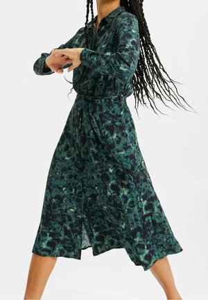 Frau mit langen Zöpfen, die ein grün gemustertes Midi-Kleid mit Knöpfen und Gürtel trägt, posiert mit verschränkten Armen und fließendem Kleid.