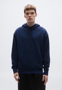 Hanorac bleumarin cu glugă, croială lejeră, prevăzut cu buzunar kangaroo, șnururi și manșete din rib. Textură netedă și branding minimalist.