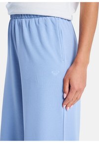 Pantalones de color azul claro, con cintura elástica, bolsillos laterales y un sutil logo en el muslo. Tejido suave y liso con un ajuste relajado.