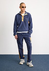 Pull en polaire marine avec des accents orange et vert, col zippé à mi-hauteur et poche frontale. Assorti avec un pantalon marine avec des détails colorés.