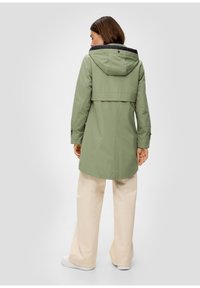 Femme portant un imperméable à capuche vert clair avec des manches longues, un pantalon large beige et des baskets blanches, de dos contre un fond uni.