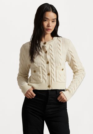 Femme portant un gilet boutonné en tricot torsadé crème avec des poches avant et un pantalon noir, se tenant avec les mains près des poches sur un fond blanc.
