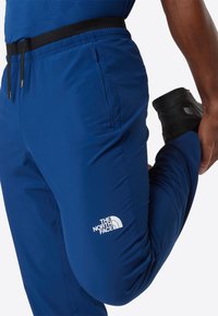 Muž oblečený v modrých kalhotách The North Face se natahuje a drží nohu za sebou, logo viditelné na stehně, bílý pozadí.