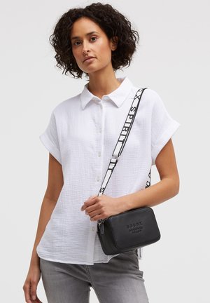 Woman with short curly hair wears a white short-sleeve button shirt and gray pants, holding a black textured crossbody bag with branded strap.