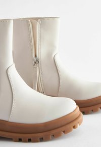 Bottines blanches en simili cuir lisse, avec une fermeture éclair sur le côté, une semelle robuste marron et des détails de couture minimalistes.