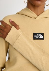 Kremowa bluza z kapturem o ribbowym wykończeniu, z czarną naszywką z logo, pokazana na ręce opierającej się na ramieniu.