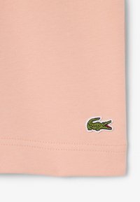 Lichtroze stof met een klein groen geborduurd krokodillenlogo nabij de gestikte zoom aan de onderkant aan de rechterzijde.