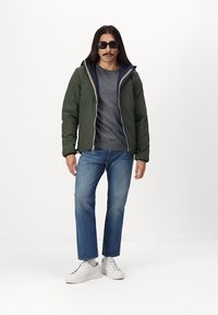 Grøn quilted jakke med marineblå hætte og gule detaljer, grå t-shirt, blå lige ben jeans og hvide sneakers. Glat tekstur.