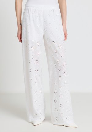 Femme portant un pantalon blanc à jambes larges avec un motif floral en broderie anglaise et une taille élastique, associé à des chaussures blanches à bout pointu.