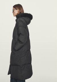 Schwarze Daunenjacke mit Kapuze, ausgestattet mit einem Kunstfellbesatz, langem Schnitt, gestepptem Design, seitlichen Taschen und einem Reißverschluss auf der Vorderseite.