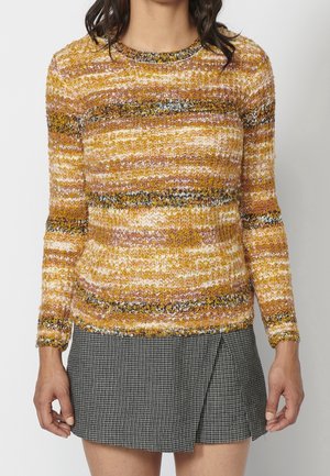 Woman wearing a long-sleeve textured sweater with horizontal mustard, pink, white, and black stripes, paired with a black-and-white houndstooth skirt.