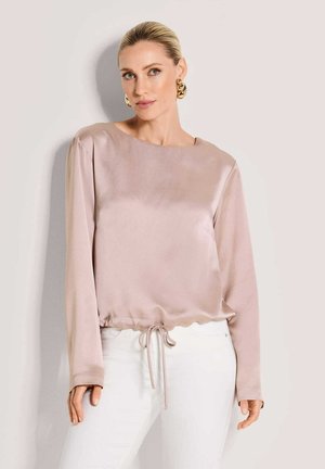 Hellrosa Satintop mit rundem Ausschnitt, langen Ärmeln und einem Saum mit Kordelzug. Glatte Textur und dezenter Glanz. Kombiniert mit weißen Hosen.
