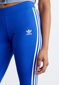 Leggings azules hechos de tela elástica con acentos de tres rayas blancas a lo largo de los lados y un logotipo de Adidas en el muslo izquierdo.