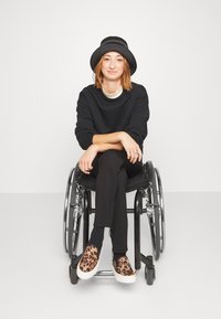 Jeune femme aux cheveux roux portant un chapeau noir et des vêtements noirs, assise en tailleur dans un fauteuil roulant, souriant à la caméra.