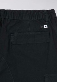 Pantaloni neri con cintura elastica, dotati di una tasca posteriore e di etichette in materiali a contrasto. La texture appare liscia con linee sottili.