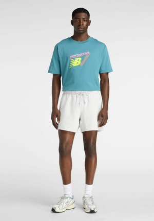 Man draagt een teal New Balance hardloop-T-shirt, lichtgrijze shorts, witte sokken en witte New Balance sneakers, staand tegen een effen achtergrond.