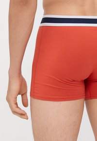 Boxer in tessuto elastico rosso con cintura a righe blu e bianche, design aderente e cuciture piatte. Mano posizionata vicino alla coscia.