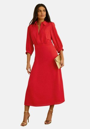 Kvinde iført en rød midi-kjole med trekvartlange ærmer og krave, der holder en guldfarvet clutch og bærer guldfarvede højhælede sandaler.
