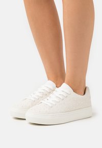 Sneaker bianche in tela con parte superiore testurizzata e piccoli dettagli iridescenti, punta rotonda e suola in gomma. I lacci sono bianchi senza elementi metallici.