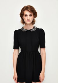 Schwarzes Kleid mit kurzen Puffärmeln und einem mit Perlen verzierten Kragen. Der Stoff ist glatt und hat vertikale Falten auf der Vorderseite.