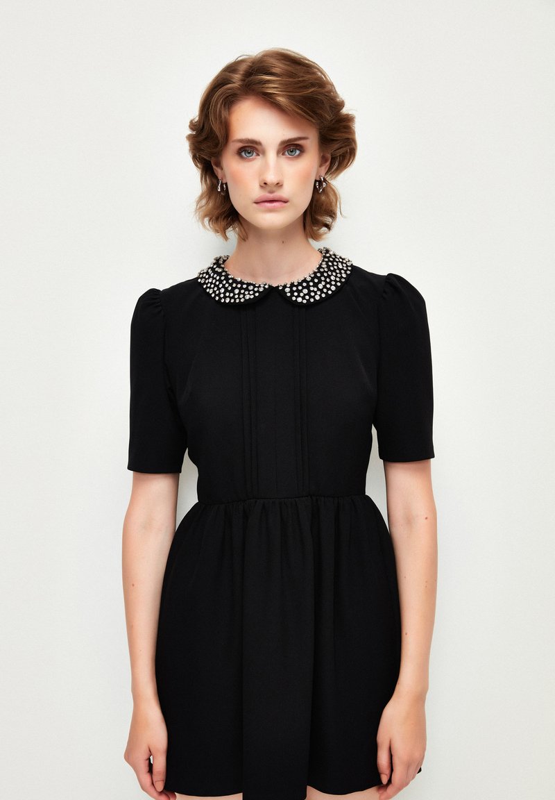 Schwarzes Kleid mit kurzen Puffärmeln und einem mit Perlen verzierten Kragen. Der Stoff ist glatt und hat vertikale Falten auf der Vorderseite.