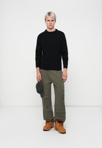 Pulover negru tricotat, pantaloni cargo verzi măslin cu buzunare, șapcă gri în mână și cizme bej. Design simplu, cu un aspect ajustat.