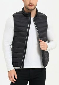 Gilet en doudoune noir avec un design matelassé horizontal, col haut et fermeture éclair intégrale. Porté par-dessus une chemise à manches longues blanche, il ne comporte pas de poches extérieures.