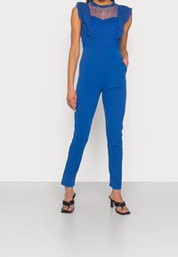Blå jumpsuit med en volangöverdel utan ärmar, spetsdetalj och åtsittande byxor. Har två sidofickor och ett mjukt, stretchigt tyg.