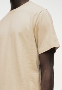 Μπεζ κοντομάνικη μπλούζα με στρογγυλή λαιμόκοψη, που φοριέται από άτομο με σκουρόχρωμο δέρμα, εμφανιζόμενο από τον λαιμό έως το μέσο του κορμού σε απλό φόντο.