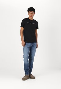 T-shirt en coton noir avec le logo "TOMMY JEANS", associé à un jean denim bleu clair et des chaussures en suede marron, le mannequin se tient contre un fond blanc.
