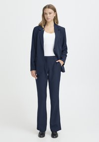 Costume bleu marine composé d'un blazer ajusté et d'un pantalon à jambes droites, tous deux en tissu lisse, avec un haut blanc en dessous.