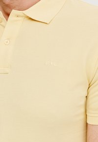 Żółta bawełniana koszulka polo o teksturowanym wykończeniu, z trzema guzikami i haftowanym logo na piersi. Krótkie rękawy.