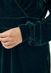 Smaragdgrünes Samtkleid mit gerüschtem Ärmelabschluss. Der Stoff hat eine weiche, glatte Textur. Es hat einen V-Ausschnitt und eine geraffte Taille.
