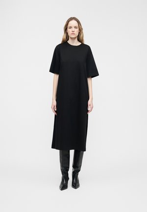 Mujer con cabello largo y rubio que lleva un vestido midi negro y holgado con mangas cortas y botas negras altas hasta la rodilla con punta, de pie frente a un fondo blanco.