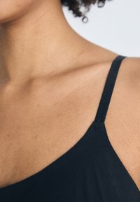 Približnji posnetek vratu in ramena osebe, ki nosi črn top s tankimi naramnicami, deloma vidni kodrasti lasje.