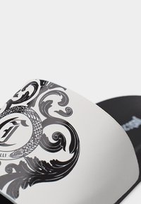 Κοντινό πλάνο λευκού σανδαλιού slide με μαύρο διακοσμητικό σχέδιο στροβιλισμού και φιδιού στο λουρί και μαύρη σόλα με λευκό λογότυπο.