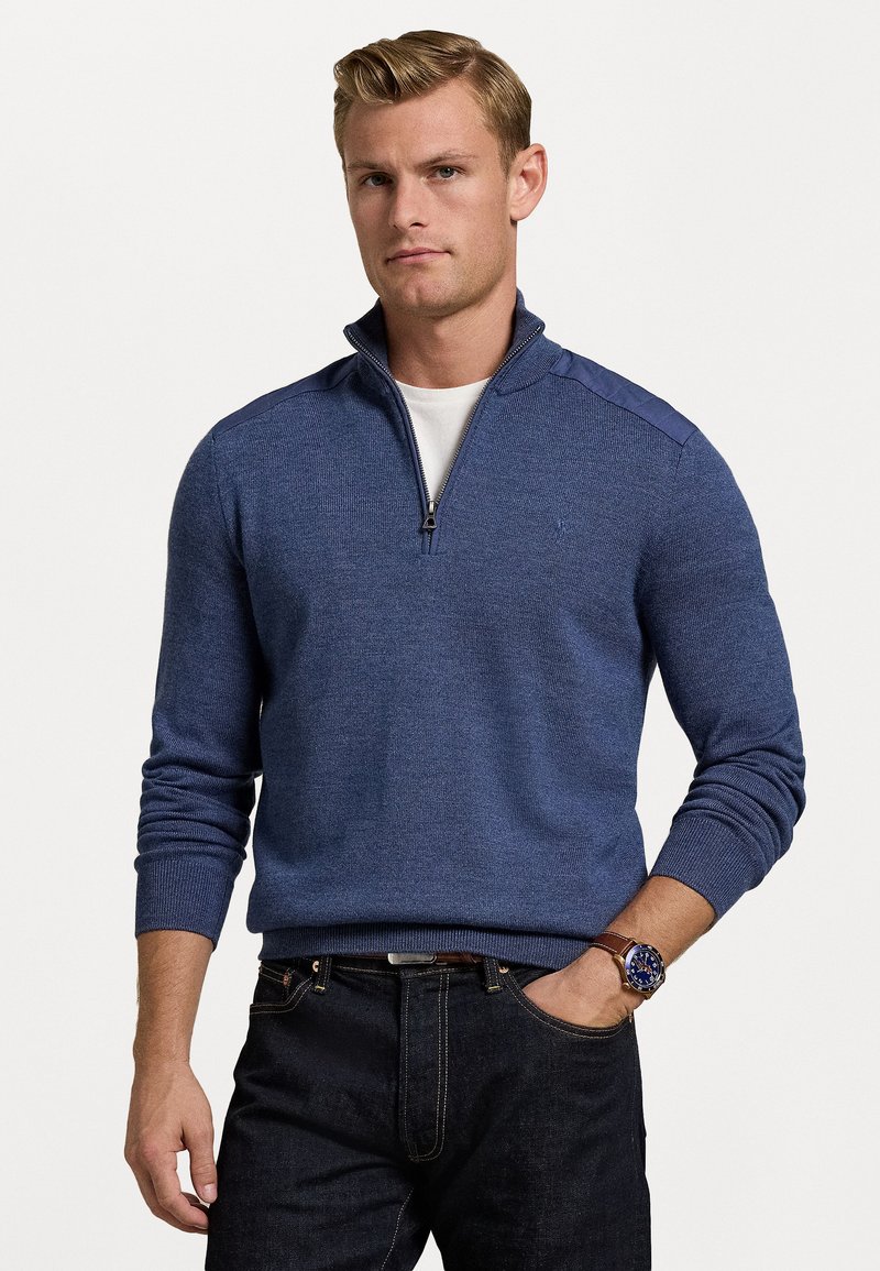Navy blue zip-up sweater with ribbed cuffs and collar, featuring a textured knit pattern. Worn over a white shirt; paired with dark jeans.