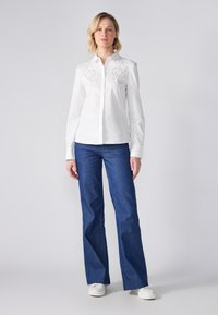 Camicia bianca con bottoni e dettagli in pizzo sulle spalle, abbinata a jeans in denim blu svasati e sneakers bianche. Texture liscia ovunque.