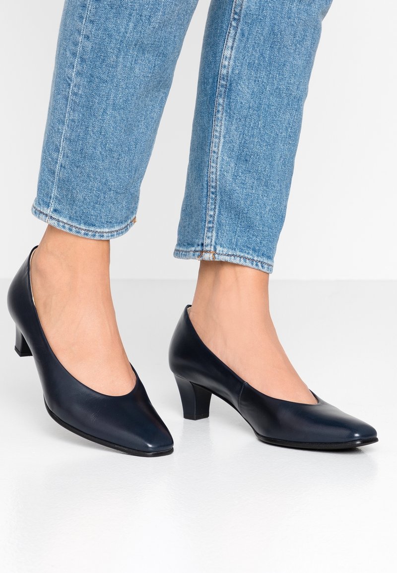 Femme portant des escarpins en cuir bleu marine à talons bas assortis à un jean denim bleu clair court, sur un sol blanc.