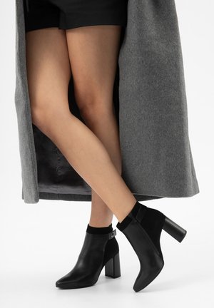 Nohy překřížené, obuté v černých kotníkových botách s širokým podpatkem, černé punčocháče a dlouhý šedý kabát rozepnutý vpředu.