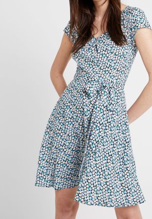 Kvinde iført en knælang kjole med kortærmer, blåt og hvidt blomsterprint og et bundet bælte i taljen.