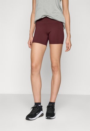 Kvinde iført vinrøde tætsiddende shorts, grå ærmeløs top, sorte Nike-sneakers og sorte ankelsokker, der står foran en ensfarvet baggrund.