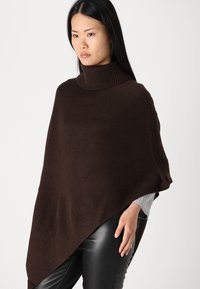 Brun strikket poncho med høy hals, med løs passform og asymmetrisk nederkant. Teksturert stoff med ribbet detaljer.