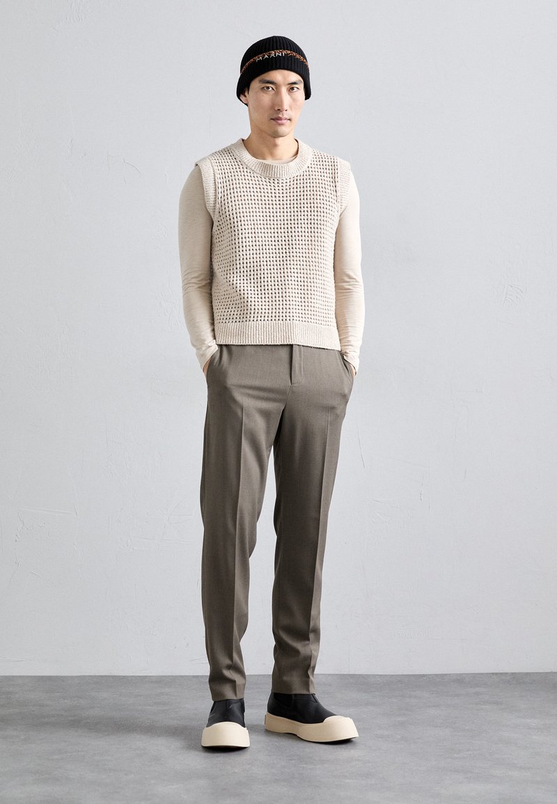 Mand iført sort beanie, beige ærmeløs strikvest over langærmet skjorte, grå bukser og sorte sko med tykke beige såler, stående med hænderne i lommen.