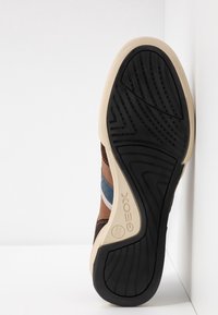 Baskets marron et bleues avec une semelle en caoutchouc texturée, une semelle intermédiaire beige lisse et une semelle extérieure à motifs. Logo de la marque visible.