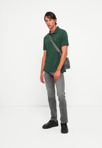 Man staande in een donkergroen poloshirt, grijze jeans, zwarte schoenen en een grijze schoudertas tegen een effen witte achtergrond.