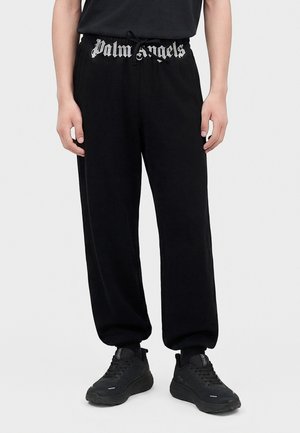Personne portant un jogging noir avec le texte "Palm Angels" sur la ceinture, des baskets noires et un t-shirt noir, debout devant un fond uni.